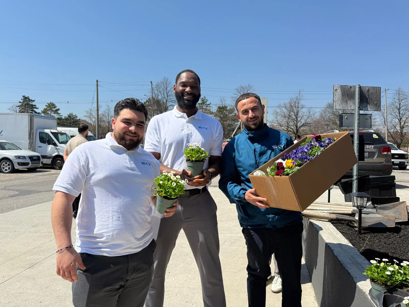 Three team members holding potted flowers and a box of seedlings beside a planter outside the office