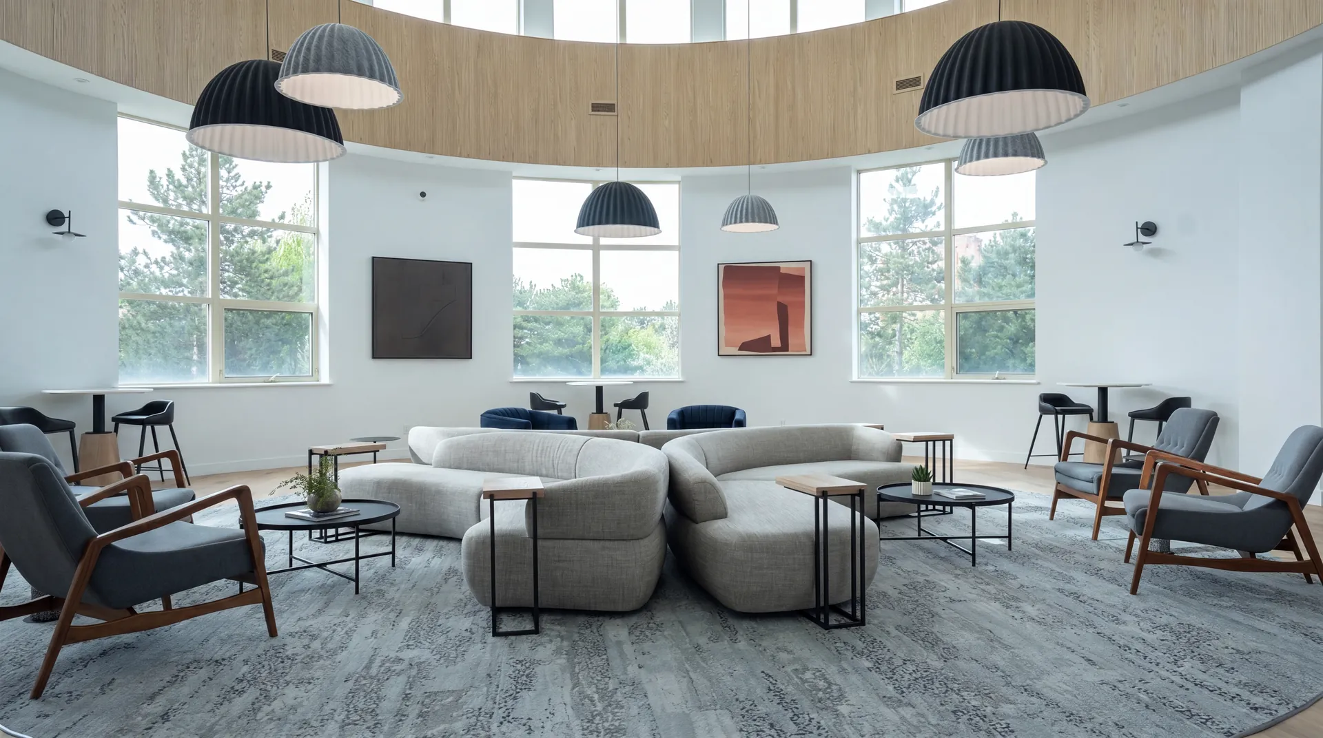 Rotunda lounge under a curved wood ceiling band, with grey modular sofas, dome pendant lights and tall windows